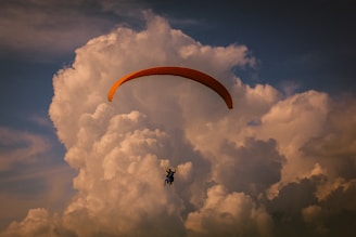 A paraglider soaring through the sky in front of a backdrop of large, dramatic clouds. The parachute is prominently shaped and colored in a striking shade that stands out against the soft, fluffy clouds and deep blue sky.