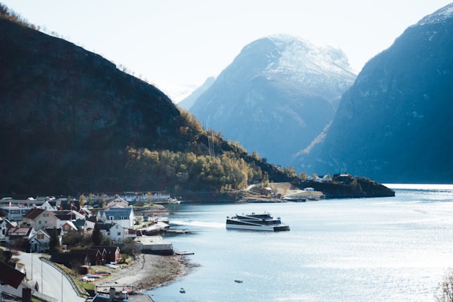 A scenic fjord landscape with a calm body of water reflecting light. A ferry is navigating the waterway surrounded by steep, mountainous terrain. A small village with colorful houses nestles at the water's edge, bordered by lush greenery. Snow-capped peaks rise in the background, adding to the serene and picturesque setting.