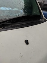 Raindrops are scattered across the windshield of a vehicle, capturing reflections of the surroundings. The wiper rests at the bottom of the windshield, and the hood appears to be a pale color with a black antenna attached.