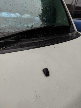 Raindrops are scattered across the windshield of a vehicle, capturing reflections of the surroundings. The wiper rests at the bottom of the windshield, and the hood appears to be a pale color with a black antenna attached.
