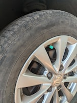 A close-up view of a car's tire and wheel, showing a silver alloy wheel with a visible logo in the center. The tire is slightly worn with visible text on the sidewall, and a small green valve cap is attached to the tire valve.