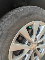 A close-up view of a car's tire and wheel, showing a silver alloy wheel with a visible logo in the center. The tire is slightly worn with visible text on the sidewall, and a small green valve cap is attached to the tire valve.