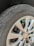 A close-up view of a car's tire and wheel, showing a silver alloy wheel with a visible logo in the center. The tire is slightly worn with visible text on the sidewall, and a small green valve cap is attached to the tire valve.