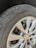 A close-up view of a car's tire and wheel, showing a silver alloy wheel with a visible logo in the center. The tire is slightly worn with visible text on the sidewall, and a small green valve cap is attached to the tire valve.