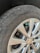 A close-up view of a car's tire and wheel, showing a silver alloy wheel with a visible logo in the center. The tire is slightly worn with visible text on the sidewall, and a small green valve cap is attached to the tire valve.