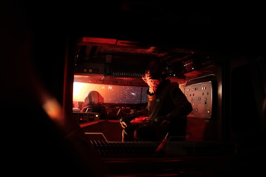 A dimly lit spaceship cockpit featuring advanced control panels and screens glowing in the darkness. The space is occupied by a figure with an alien-like appearance, wearing a suit and facing forward. The windows reveal a view of space with stars and a distant celestial body.