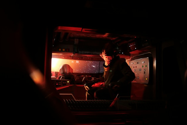 A dimly lit spaceship cockpit featuring advanced control panels and screens glowing in the darkness. The space is occupied by a figure with an alien-like appearance, wearing a suit and facing forward. The windows reveal a view of space with stars and a distant celestial body.