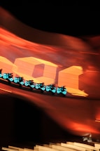 A group of futuristic motorcycles with glowing blue lights speed along a curved track. The background features large, bold orange shapes and numbers, creating a vibrant and dynamic scene with a sense of motion and excitement.