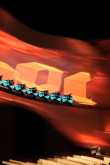 A group of futuristic motorcycles with glowing blue lights speed along a curved track. The background features large, bold orange shapes and numbers, creating a vibrant and dynamic scene with a sense of motion and excitement.