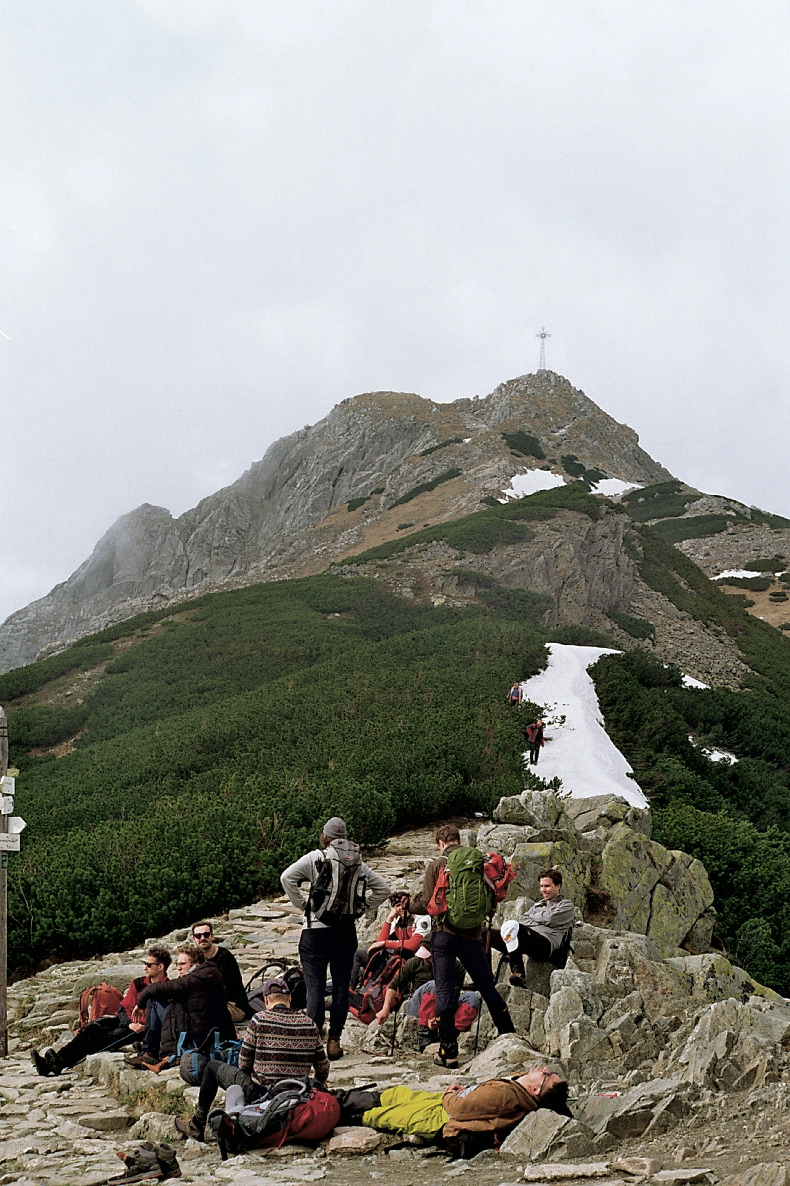 Hikers rest on a rocky mountain path near snowy slopes.
