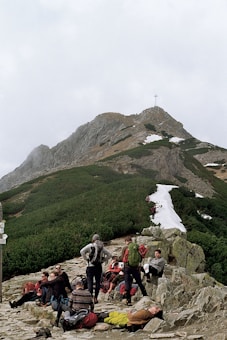 A group of hikers is resting on a rocky path in a mountainous landscape. Some are sitting, and others are standing, with backpacks and gear scattered around them. The background features a rugged green and brown mountain slope partially covered with patches of snow. A cross is visible at the mountain peak, and the sky is overcast.