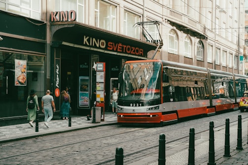 A modern red and gray tram is approaching a stop in front of a cinema named 'Kino Světozor' on a city street. Several pedestrians are walking or standing near the tram, while buildings with large windows and various advertisements surround the area. The street and tram tracks are lined with black bollards.