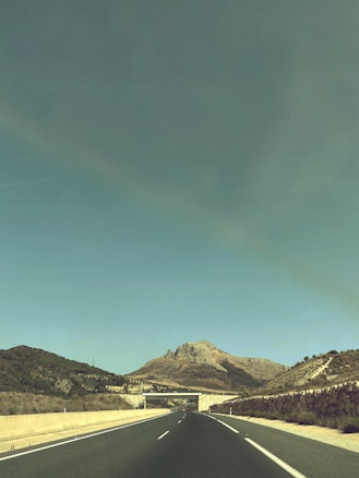 A highway stretches into the distance, leading towards a mountainous landscape under a clear blue sky. The road is flanked by low, grassy hills and sparse vegetation. A concrete overpass crosses the highway, enhancing the sense of perspective and depth.