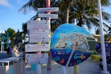 A colorful signpost stands on a sandy beach under palm trees. The sign holds several directional signs pointing to various locations such as restaurants, water parks, beaches, and other attractions. Next to it is a large, circular map illustrating the layout of an island resort called Coco Cay. People are visible in the background enjoying the area.