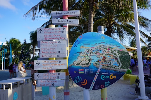 A colorful signpost stands on a sandy beach under palm trees. The sign holds several directional signs pointing to various locations such as restaurants, water parks, beaches, and other attractions. Next to it is a large, circular map illustrating the layout of an island resort called Coco Cay. People are visible in the background enjoying the area.