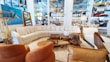 A stylish living room setup in a showroom with a large, L-shaped beige sectional sofa surrounded by various decorative items. Brown leather chairs are positioned nearby, complemented by a coffee table set displaying ornaments and a potted plant. The walls are adorned with an abstract painting and shelves stocked with colorful ceramics and glassware.