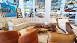 A stylish living room setup in a showroom with a large, L-shaped beige sectional sofa surrounded by various decorative items. Brown leather chairs are positioned nearby, complemented by a coffee table set displaying ornaments and a potted plant. The walls are adorned with an abstract painting and shelves stocked with colorful ceramics and glassware.