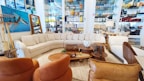 A stylish living room setup in a showroom with a large, L-shaped beige sectional sofa surrounded by various decorative items. Brown leather chairs are positioned nearby, complemented by a coffee table set displaying ornaments and a potted plant. The walls are adorned with an abstract painting and shelves stocked with colorful ceramics and glassware.