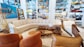 A stylish living room setup in a showroom with a large, L-shaped beige sectional sofa surrounded by various decorative items. Brown leather chairs are positioned nearby, complemented by a coffee table set displaying ornaments and a potted plant. The walls are adorned with an abstract painting and shelves stocked with colorful ceramics and glassware.