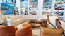A stylish living room setup in a showroom with a large, L-shaped beige sectional sofa surrounded by various decorative items. Brown leather chairs are positioned nearby, complemented by a coffee table set displaying ornaments and a potted plant. The walls are adorned with an abstract painting and shelves stocked with colorful ceramics and glassware.