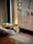 A small dog is laying on a plush, circular bed next to a large sliding glass door. The room is illuminated with natural light filtering through the glass, highlighting the wooden floor and the cozy atmosphere. Outside, a blurred view of chairs and a deck is visible.