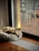 A small dog is laying on a plush, circular bed next to a large sliding glass door. The room is illuminated with natural light filtering through the glass, highlighting the wooden floor and the cozy atmosphere. Outside, a blurred view of chairs and a deck is visible.