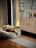 A small dog is laying on a plush, circular bed next to a large sliding glass door. The room is illuminated with natural light filtering through the glass, highlighting the wooden floor and the cozy atmosphere. Outside, a blurred view of chairs and a deck is visible.