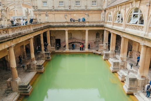 An ancient Roman-style bath surrounded by columns and stone statues, with weathered stone architecture. The pool is filled with greenish water and several people are scattered around, some observing from a higher walkway.