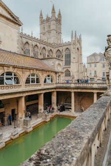 An ancient architectural structure featuring a large, open bathing area surrounded by stone columns and archways. In the background, a historic church with detailed stonework and a tall tower is visible. Several people are present around the bathing area, adding a sense of activity and historical tourism.