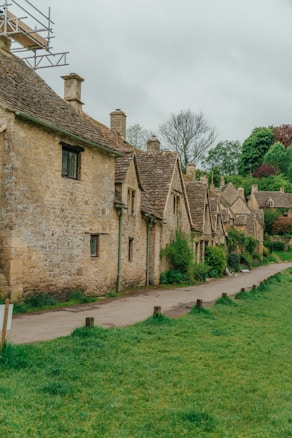 A row of rustic stone cottages lines a narrow path, surrounded by lush greenery. The architecture is traditional, characterized by steeply pitched roofs and small, rectangular windows. The foreground features a grassy area, while the background is dominated by trees under a cloudy sky.