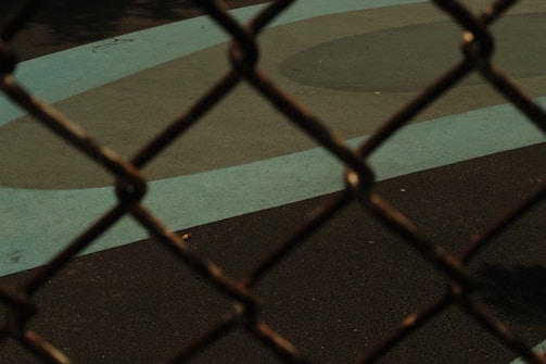 A metal chain-link fence with a blurred focus in the foreground, set against a playground or sports court surface. The ground features geometric patterns in different shades of blue and gray.