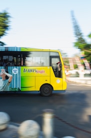 A yellow public transit bus is captured in motion on a sunny day, with blurred background elements indicating speed. The bus has the logo 'trans jogja' prominently displayed on its side, along with an advertisement featuring a person shielding their eyes. The road appears to be in an urban setting with some greenery visible.
