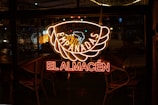 A neon sign displaying the words 'Empanadas El Almacén' is glowing against a window. The light is reflected on the glass, with raindrops visible on the window. Chairs and tables inside the establishment can be seen in the dimly lit interior.
