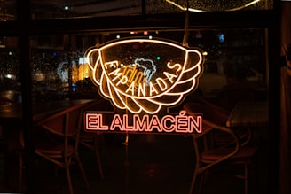 A neon sign displaying the words 'Empanadas El Almacén' is glowing against a window. The light is reflected on the glass, with raindrops visible on the window. Chairs and tables inside the establishment can be seen in the dimly lit interior.
