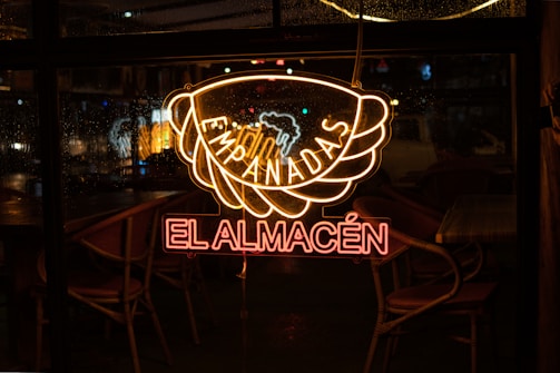 A neon sign displaying the words 'Empanadas El Almacén' is glowing against a window. The light is reflected on the glass, with raindrops visible on the window. Chairs and tables inside the establishment can be seen in the dimly lit interior.