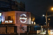 A nighttime scene featuring a commercial building with illuminated signs for 'Selva Negra' and 'Helados Artesanales.' The area is dimly lit by streetlights, with closed shutters on the ground level. A sidewalk runs alongside the building and streetlights line the road, casting a warm glow on the wet pavement.