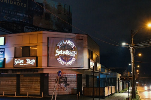 A nighttime scene featuring a commercial building with illuminated signs for 'Selva Negra' and 'Helados Artesanales.' The area is dimly lit by streetlights, with closed shutters on the ground level. A sidewalk runs alongside the building and streetlights line the road, casting a warm glow on the wet pavement.