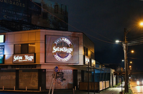 A nighttime scene featuring a commercial building with illuminated signs for 'Selva Negra' and 'Helados Artesanales.' The area is dimly lit by streetlights, with closed shutters on the ground level. A sidewalk runs alongside the building and streetlights line the road, casting a warm glow on the wet pavement.