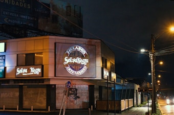 A nighttime scene featuring a commercial building with illuminated signs for 'Selva Negra' and 'Helados Artesanales.' The area is dimly lit by streetlights, with closed shutters on the ground level. A sidewalk runs alongside the building and streetlights line the road, casting a warm glow on the wet pavement.