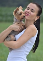 A woman holding a small dog while making a playful and affectionate face toward it. The woman is wearing a white sleeveless top and gold hoop earrings. The small dog appears to be a Yorkshire Terrier with a light brown coat and wearing a collar. The background is a blurred green field, which suggests the scene is set outdoors.
