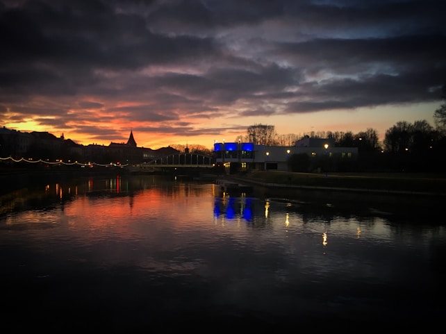 A serene view of a river reflecting the vibrant hues of a sunset sky. The horizon is adorned with silhouettes of buildings, one with a distinct pointed roof. A bridge with string lights spans across the river, while trees and a modern building with blue lights stand in the background.