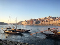 Traditional boats are floating on a wide river as the sun sets, casting a warm glow over a cityscape with densely packed, colorful buildings climbing up a hill. The foreground shows stone paving along the riverside.