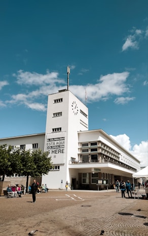 A modernist white building with rectangular windows and a clock on the tower. A large banner reads 'EL COLONIALISMO ESTÁ AQUÍ COLONIALISM IS HERE.' People are gathered in the courtyard, some sitting on benches and others walking. The sky is bright blue with fluffy clouds.