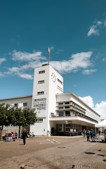 A modernist white building with rectangular windows and a clock on the tower. A large banner reads 'EL COLONIALISMO EST&Aacute; AQU&Iacute; COLONIALISM IS HERE.' People are gathered in the courtyard, some sitting on benches and others walking. The sky is bright blue with fluffy clouds.