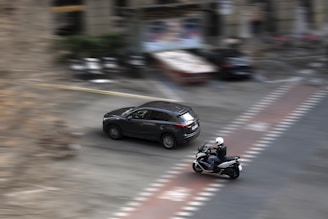 A motorcycle and a car are seen moving at speed on a city street. The motorcycle is slightly ahead of the car, with both vehicles creating a blur effect due to their motion. The surroundings are urban with blurred buildings in the background, indicating a sense of motion and speed.