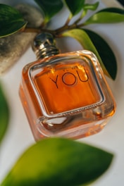 A square-shaped perfume bottle with amber-colored liquid is placed on a light surface, surrounded by green leaves and a textured gray stone. The bottle has 'YOU' prominently displayed on the front, suggesting a luxurious fragrance.
