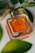 A square-shaped perfume bottle with amber-colored liquid is placed on a light surface, surrounded by green leaves and a textured gray stone. The bottle has 'YOU' prominently displayed on the front, suggesting a luxurious fragrance.