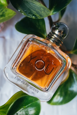 A square glass perfume bottle with the label 'YOU' and 'EMPORIO ARMANI' is surrounded by lush green leaves. The amber liquid inside the bottle contrasts with the vibrant green foliage, and there are water droplets visible on the surface of the bottle, adding a fresh and natural aesthetic.