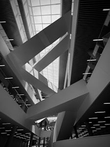 A modern architectural interior with dramatic angular lines and a geometric design. The ceiling features a large skylight allowing natural light to illuminate the space. The structure has multiple levels with sleek staircases or ramps, and the building materials appear to be metal and glass.