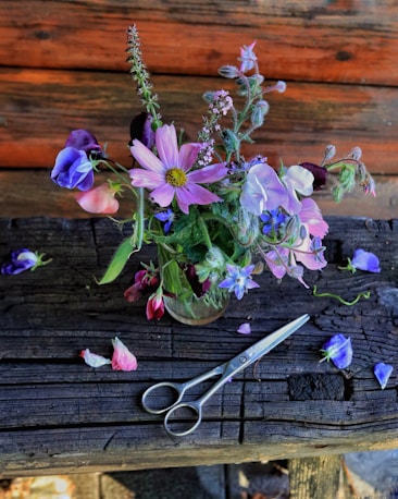 A rustic wooden table with a small glass vase holding a vibrant arrangement of wildflowers, including pink, purple, and blue blossoms. Scattered flower petals and a pair of metal scissors lie on the table, adding to the natural, handcrafted setting. The wooden background enhances the earthy, cozy feel of the scene.